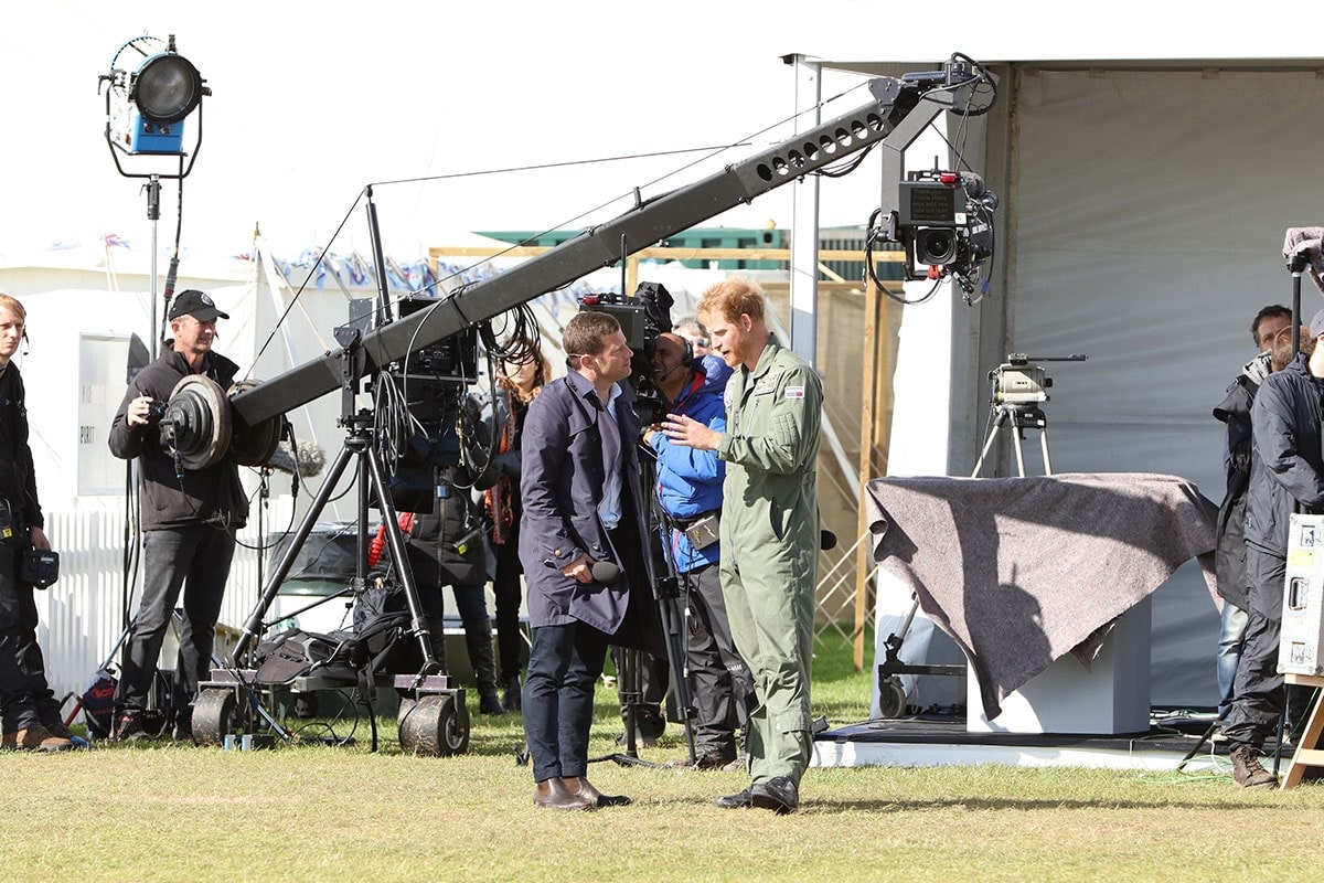 Prince Harry in a green flight suit speaks to another man holding a microphone in an outdoor setting, surrounded by a film crew with cameras and equipment.